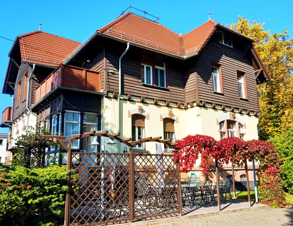 Die Villa Rochsburg mit herbstlich gefärbtem Wein an einer Pergola vor dem zweigeschossigen Gebäude mit Fachwerkobergeschoss und rotem Ziegeldach